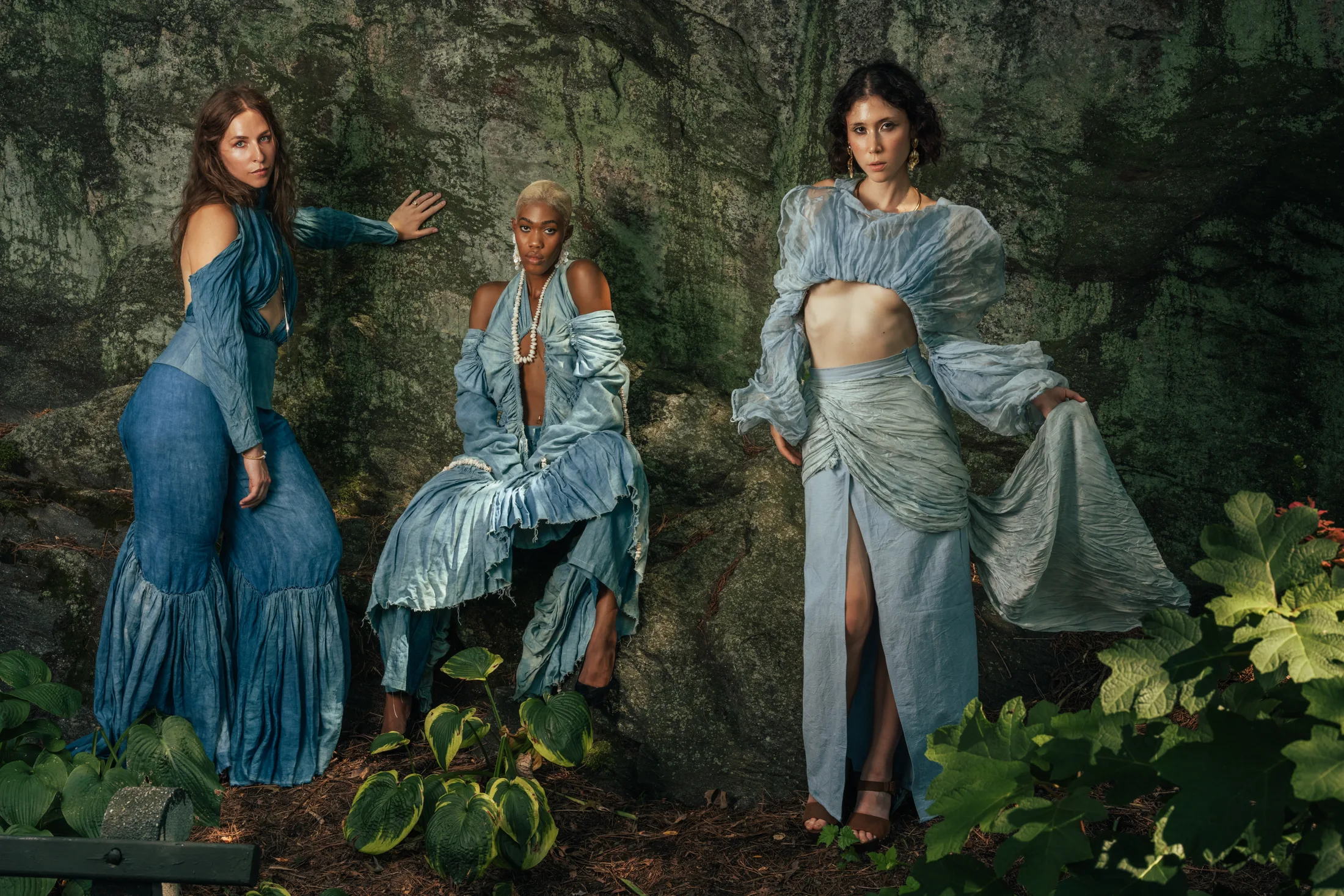 Three models in hand-dyed blue garments against a mossy rock wall