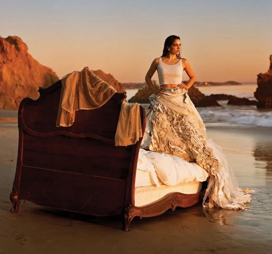 Rosalía in a ruffled ivory skirt standing on a bed on the beach at sunset